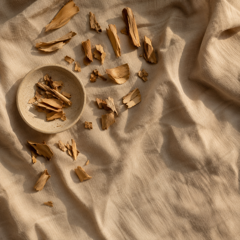 Raw sandalwood heartwood chips scattered across rumpled oatmeal linen with a small stoneware dish, seen from above in warm evening light