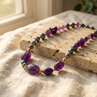 Purple Amethyst Chunky Necklace with 18k gold accents and dark pearls draped over textured travertine stone next to cream linen